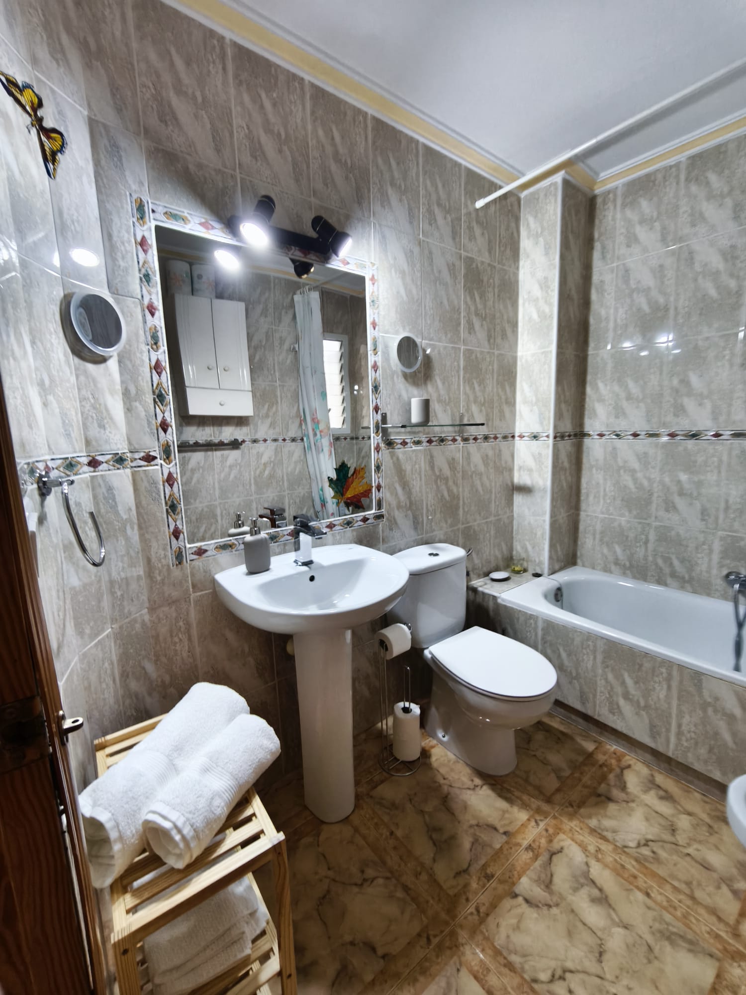 Bathroom with bathtub, sink, toilet, decorative tiled mirror frame, towel rack with fresh towels, and marble floor tiles