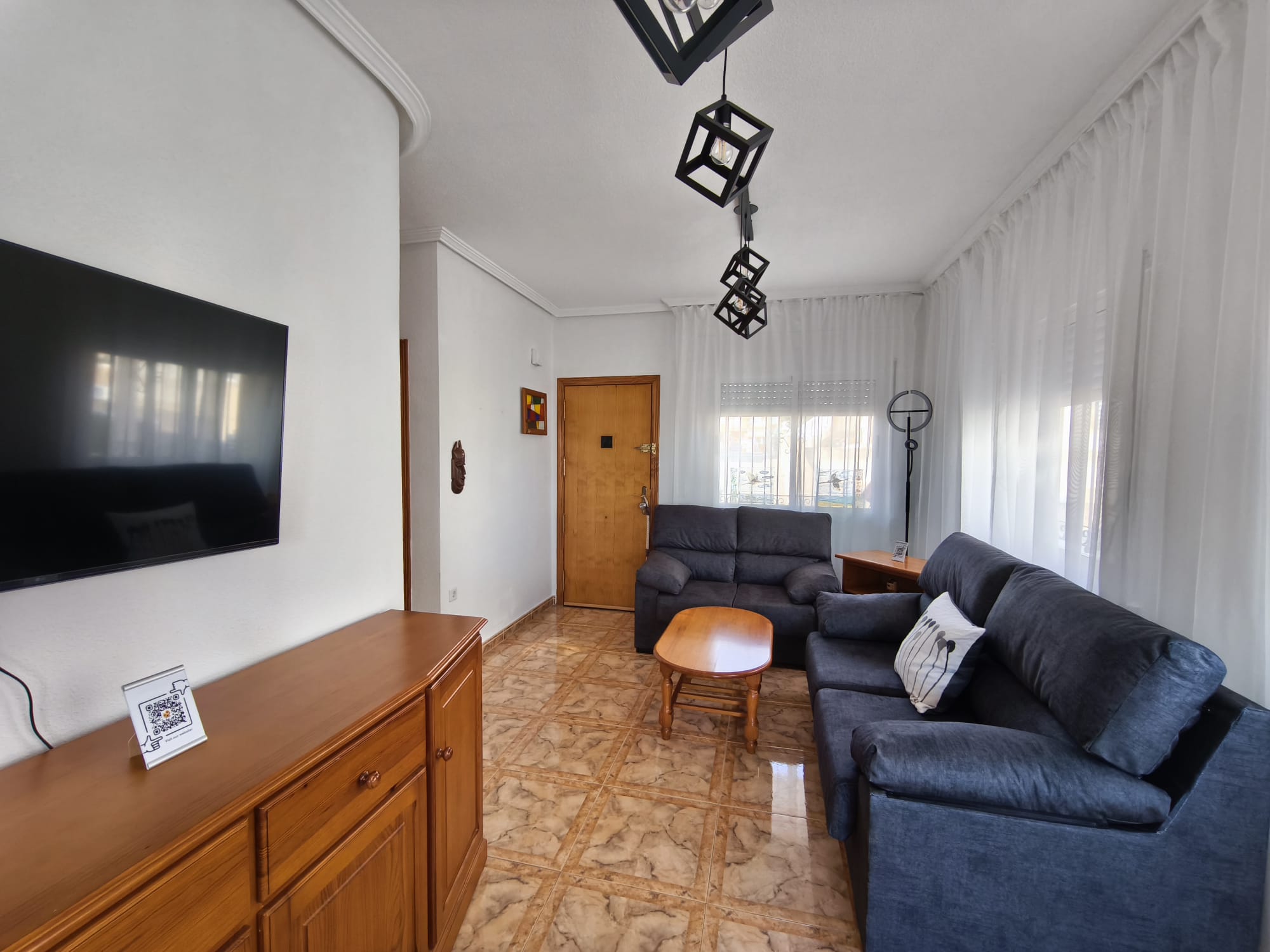 Living room with large wall-mounted TV, wooden sideboard, two dark sofas, coffee table, geometric pendant lights, and sheer curtains