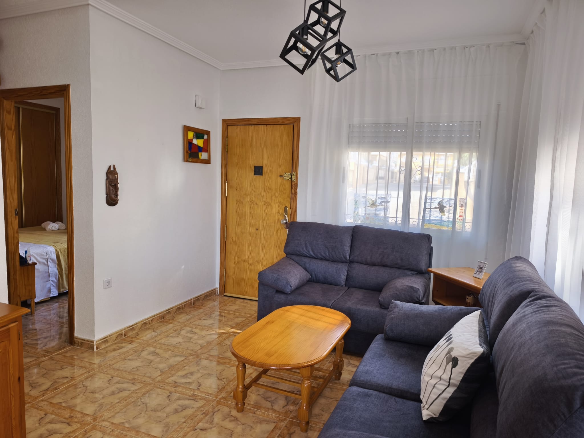 Living room with two blue sofas, wooden coffee table, geometric pendant lights, wooden front door, stained glass wall art, and a glimpse into a bedroom through an open doorway