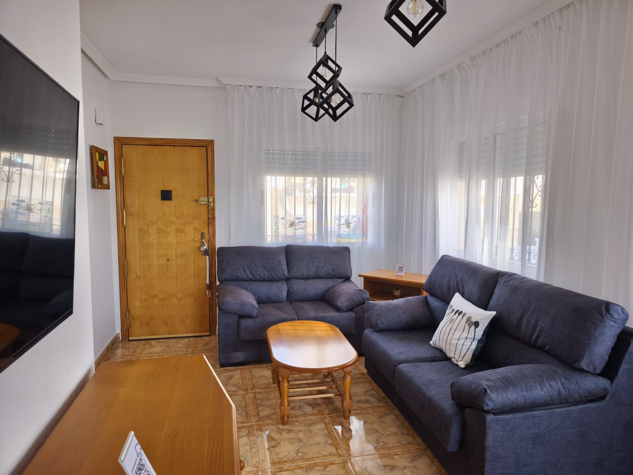 Bright living room with two blue sofas, wooden coffee table, large wall-mounted TV on wooden sideboard, geometric pendant lights, wooden front door, stained glass wall art, and sheer curtains with natural light
