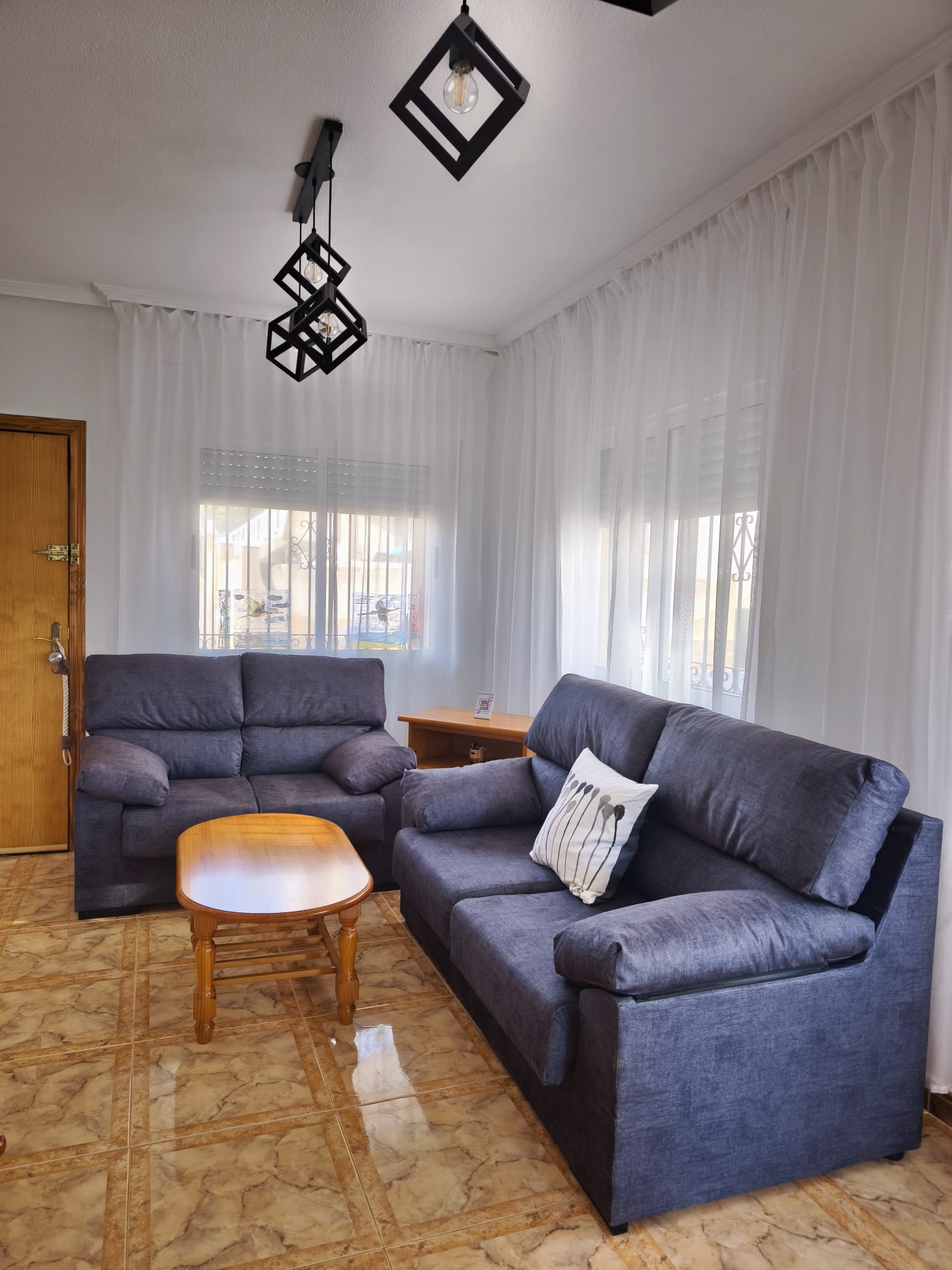 Living room corner with two blue sofas, wooden coffee table, geometric pendant lights, wooden front door, sheer curtains with natural light coming through windows with security grilles
