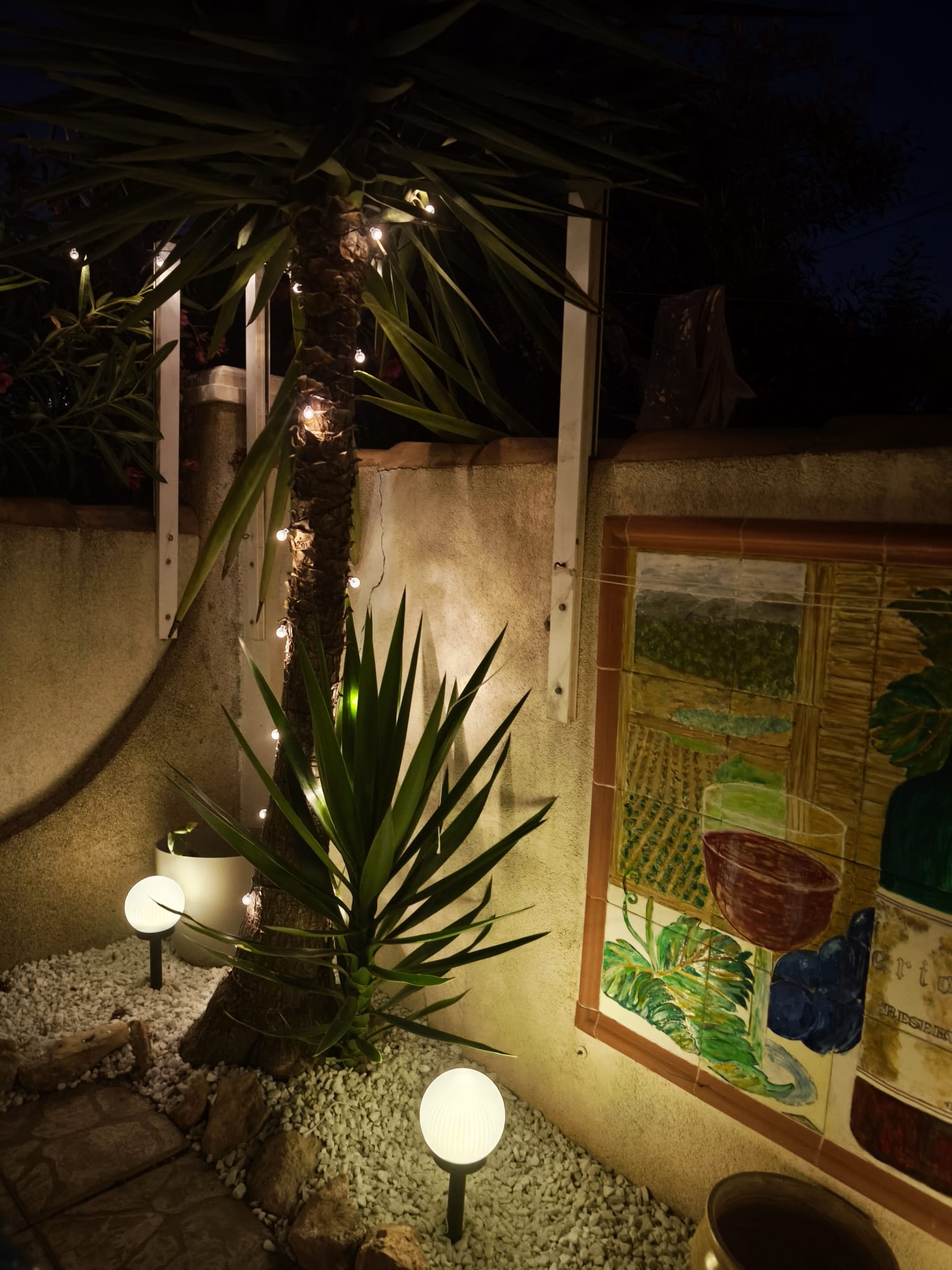 Evening garden ambiance with fairy lights on yucca palm, globe lanterns, and decorative tile mural